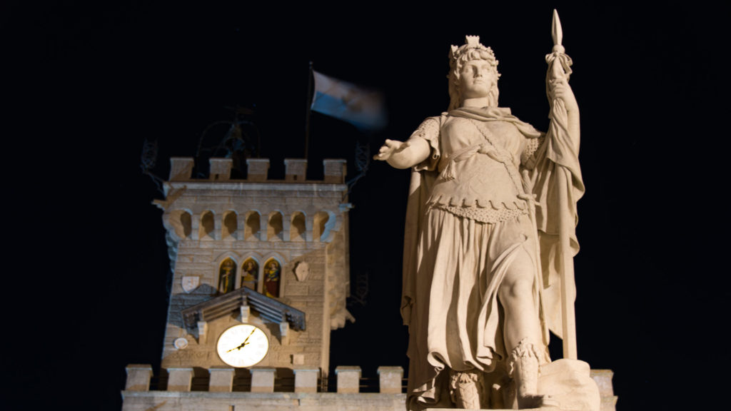 Low-angle night view of San Marino’s crowned Statue of Liberty holding a spear, with Palazzo Pubblico clock tower behind.