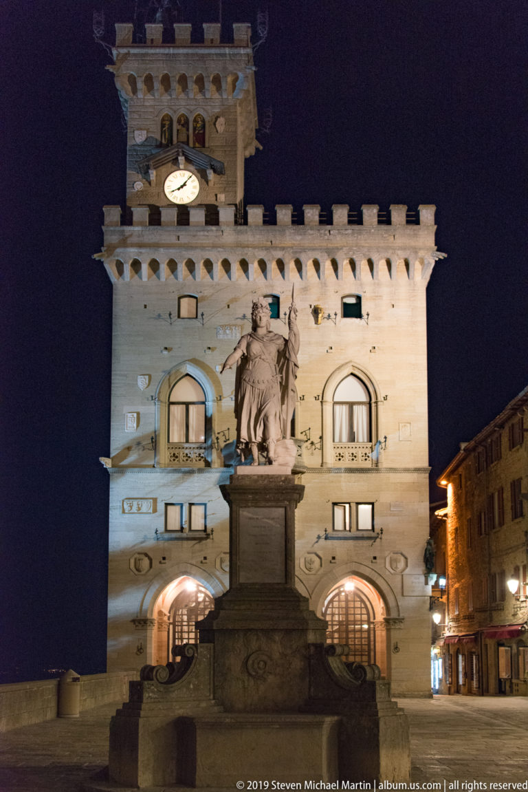 Illuminated Palazzo Pubblico with crenellations and clock tower behind the Statue of Liberty in Piazza della Libertà.