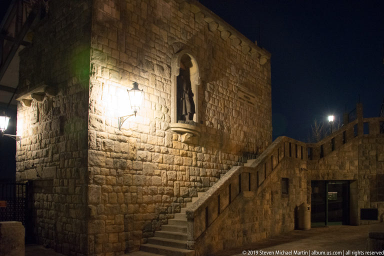 Stone stair climbing beside a rough masonry wall with an arched statue niche and warm streetlamps at night.