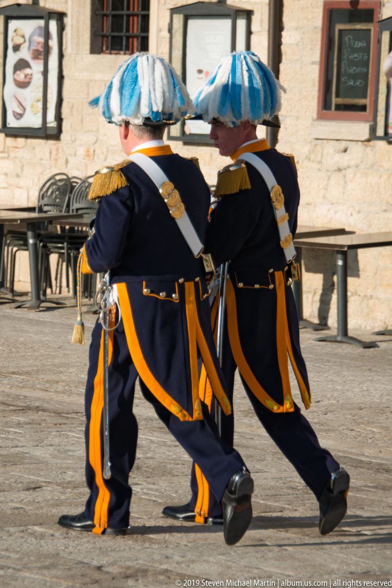 Two San Marino ceremonial guards in dark uniforms and blue-and-white plumed hats march side by side across a sunlit square.