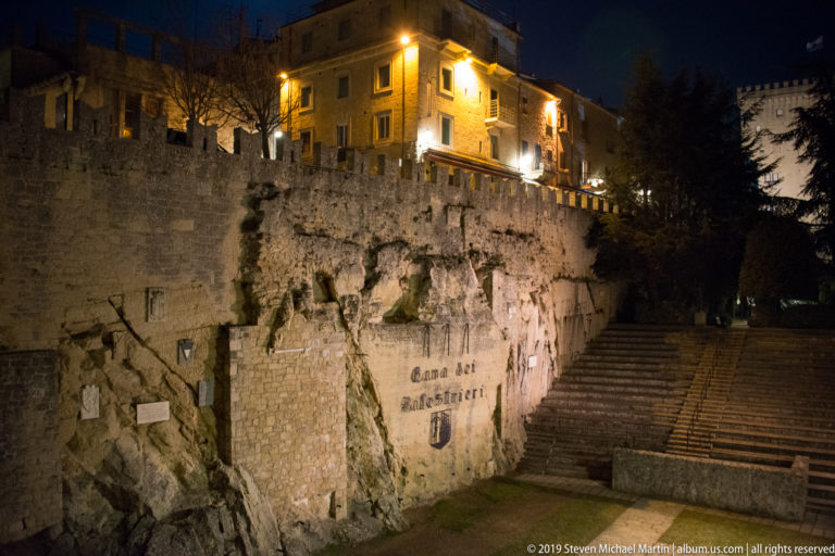 Illuminated rock face marked Cava dei Balestrieri below buildings and crenellations, with a broad stair climbing at right.