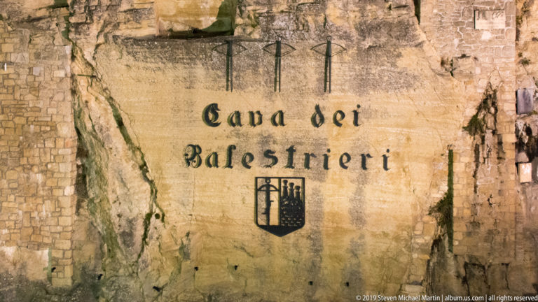 Pale rock face inscribed Cava dei Balestrieri in black Gothic lettering with a shield emblem below.