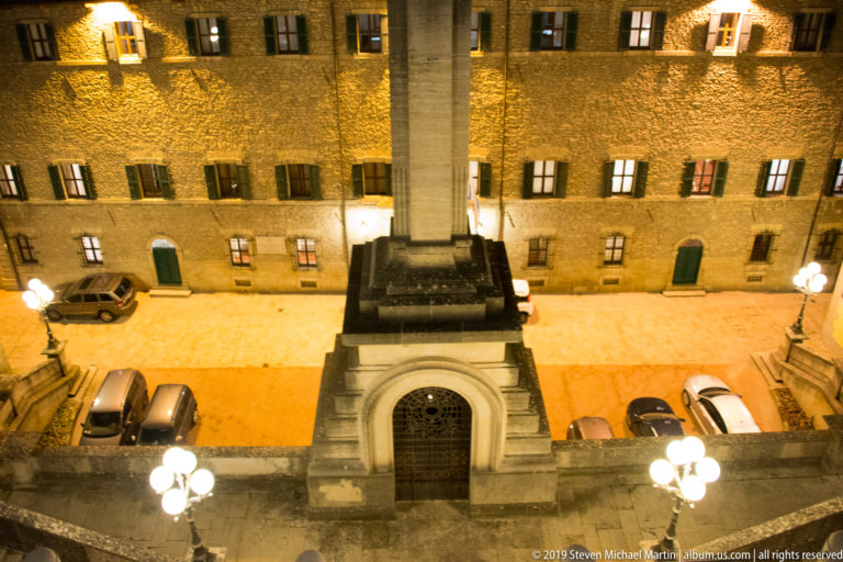 High-angle night view of a tall rectangular stone monument above an arched doorway, with parked cars and globe streetlamps.