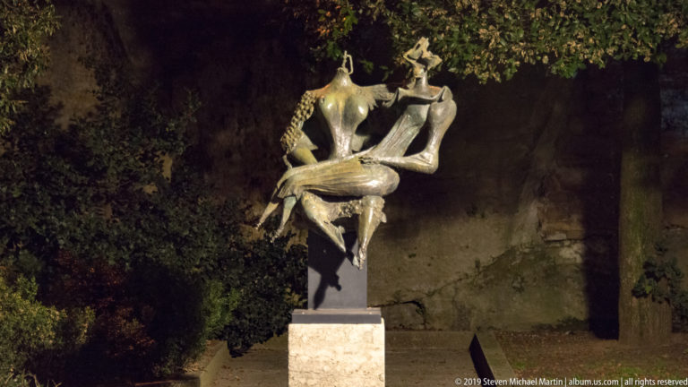 Illuminated bronze seated figure in a twisting pose on a tall stone pedestal against dark trees.