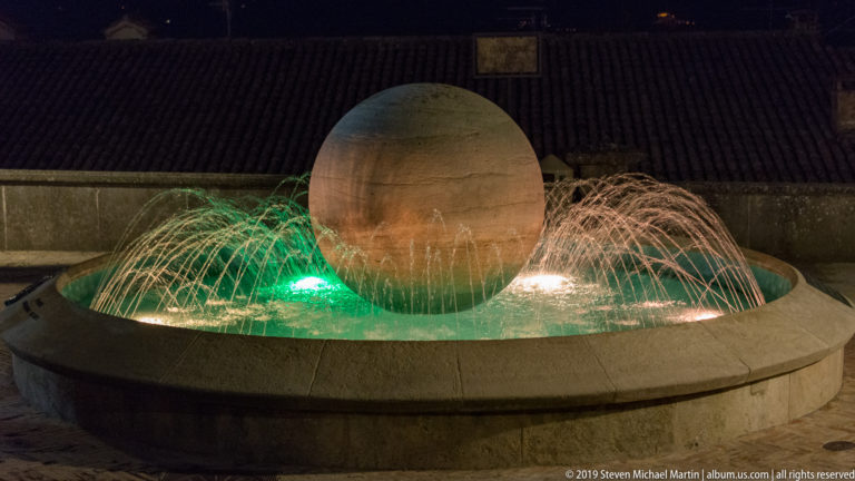 Large stone sphere centered in a circular basin with green and white illuminated water jets at night.