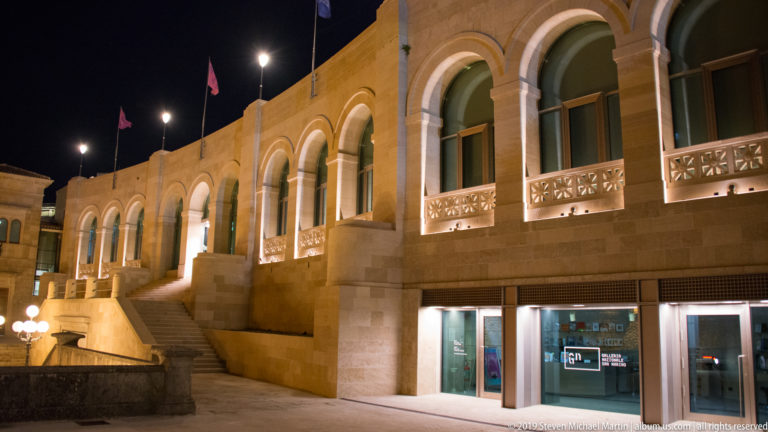 Long illuminated stone building with repeated upper arches, decorative balustrades, flags, staircase, and glazed storefronts below.