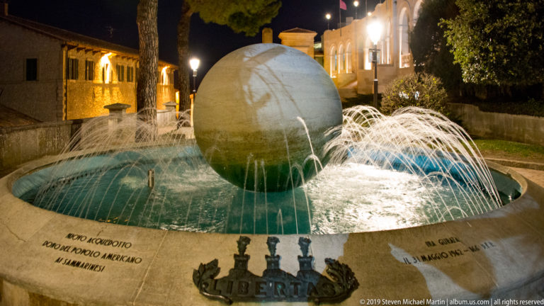 Large stone sphere in a circular fountain surrounded by arcing water jets, with LIBERTAS and three-tower emblem on the rim.