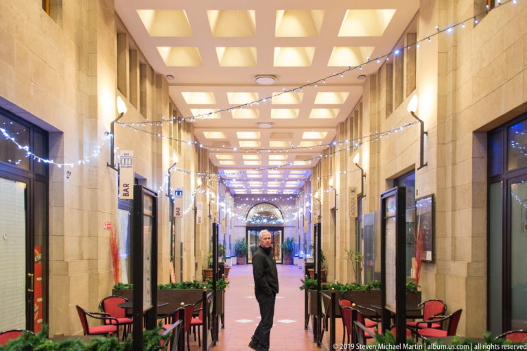 Long covered passage with glowing coffered ceiling, string lights, café tables, shopfronts, and one man standing centrally.