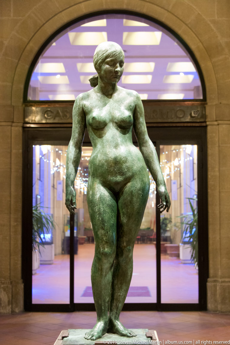 Green-patinated bronze female nude standing before a brightly lit arched glass entrance.