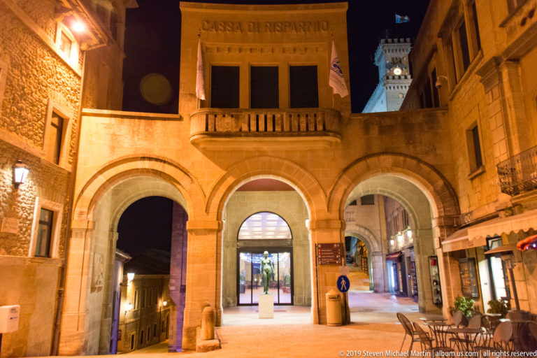 Illuminated Cassa di Risparmio façade with three arches, balcony, pedestrian passages, and a distant tower at night.
