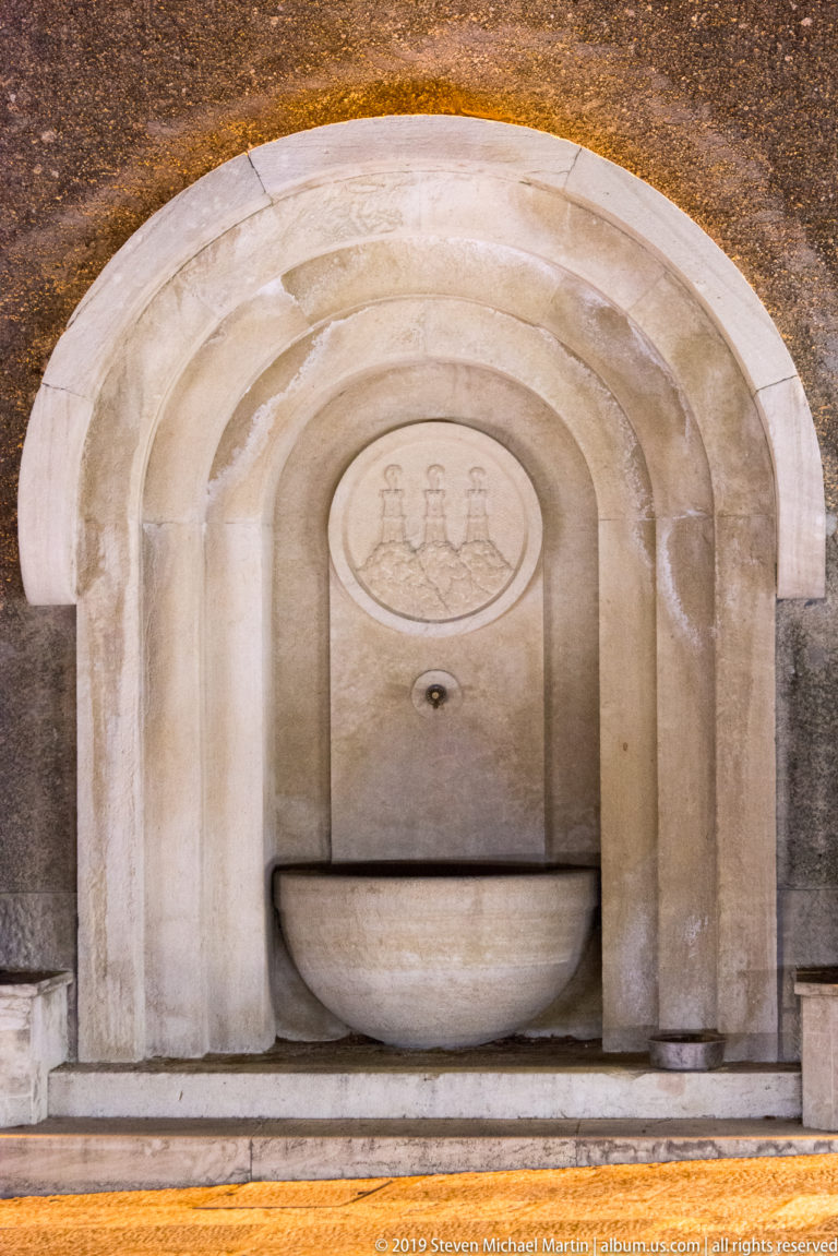 Semicircular stone wall fountain recessed in a molded niche, with a three-tower emblem above the central spout.
