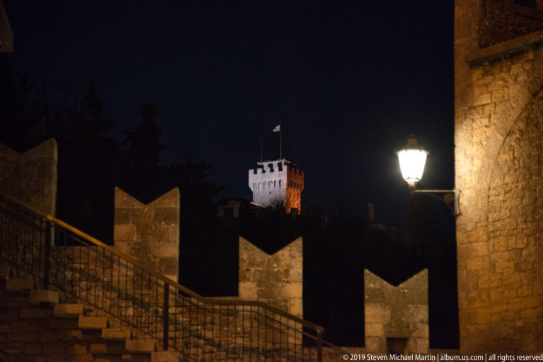 Distant illuminated fortress tower above dark crenellated walls and a stone stair, with a bright streetlamp at right.