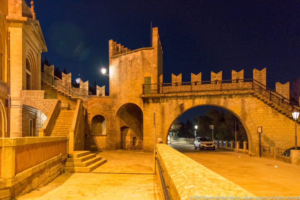 Illuminated stone gateway with broad arch, crenellated parapet, stairs, smaller arches, and square tower at night.