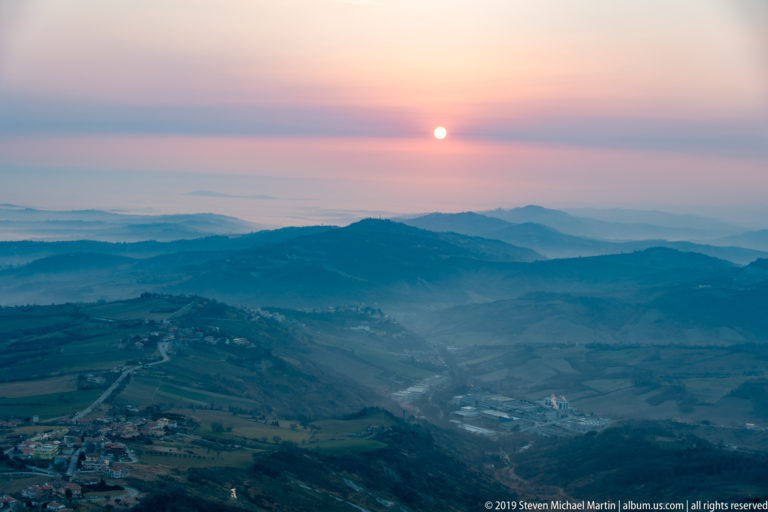 Bright setting sun in a pink sky above layered blue mountain ridges, valleys, and faint settlements.