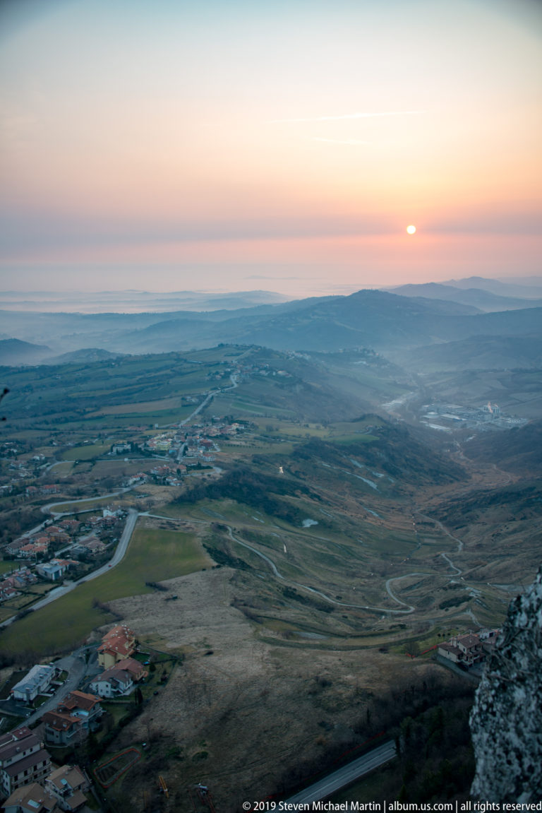 Low sun at the horizon above an open plateau with winding roads, scattered buildings, and distant ridges.