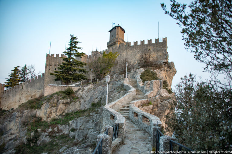 Stone stair climbing over rock toward the crenellated walls and tall keep of the Second Tower, Cesta / Fratta.