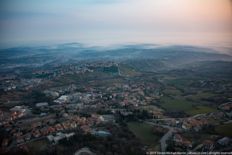 Wide view over red-roofed neighborhoods, fields, roads, and rolling countryside fading into haze.