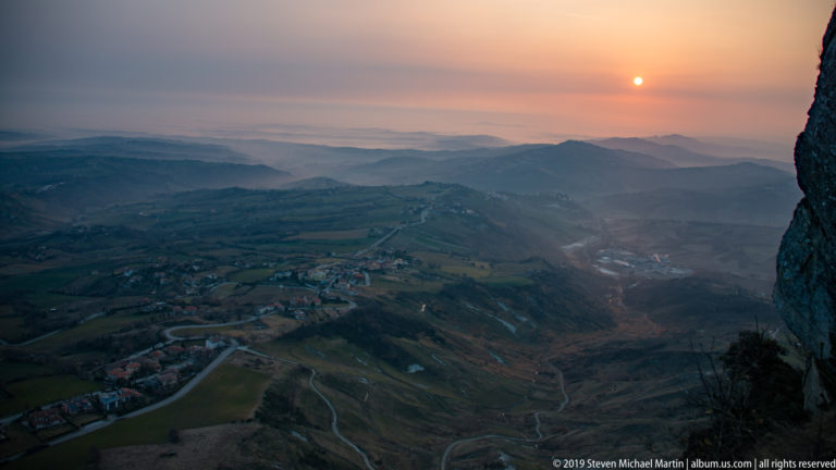 Sun setting over layered ridges, winding roads, valleys, and scattered settlements, with a dark rock face at right.