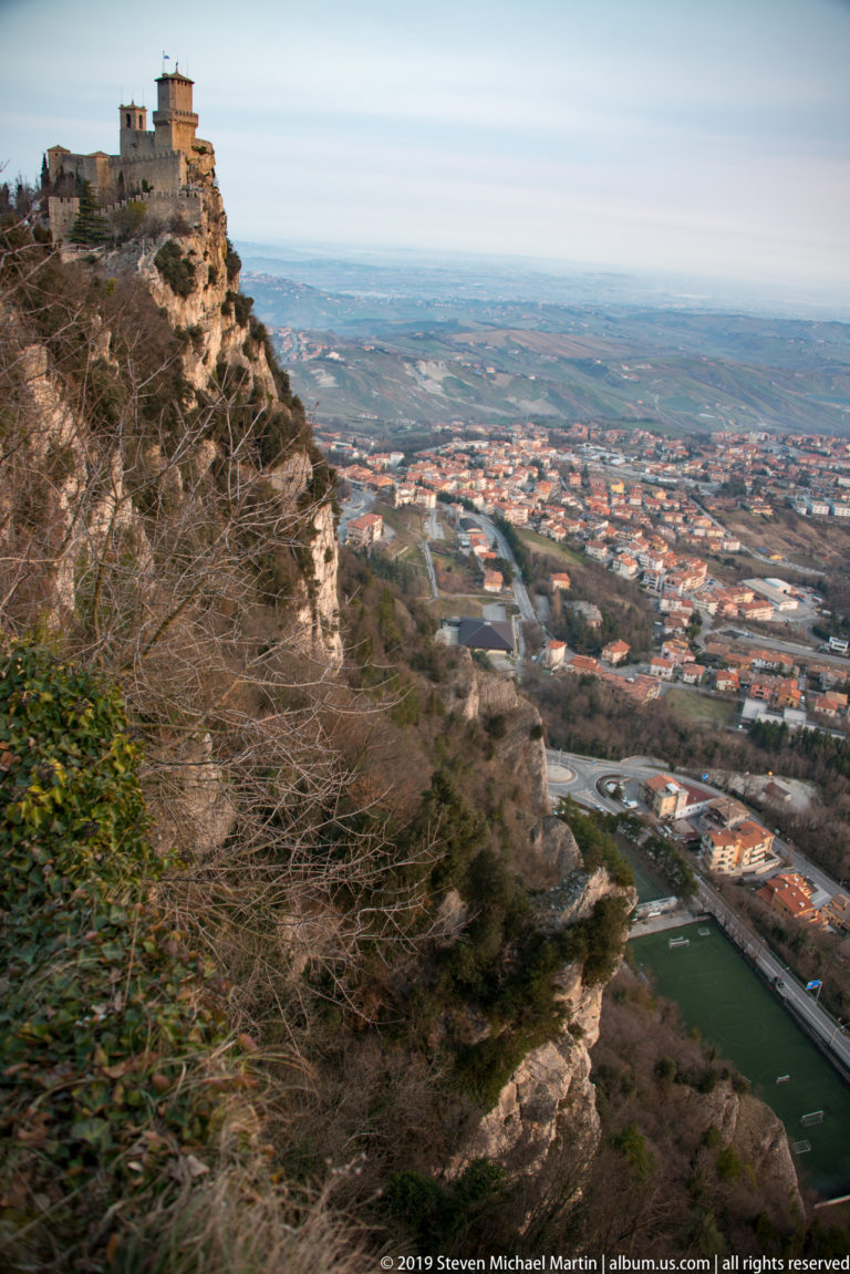 Rocky wooded slope descending toward a broad townscape, roads, fields, and distant countryside below Monte Titano.