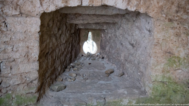 Narrow rough-stone passage with rubble on the floor leading to a bright keyhole-shaped opening.