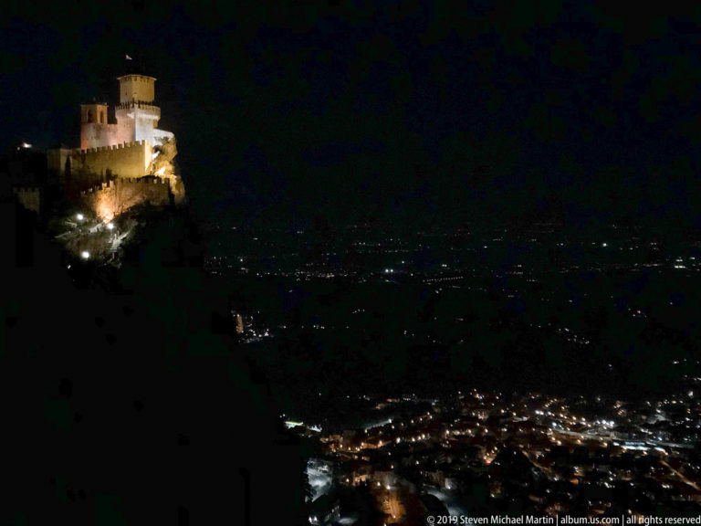 Floodlit stone fortress high on a dark ridge above clusters of city lights at night.
