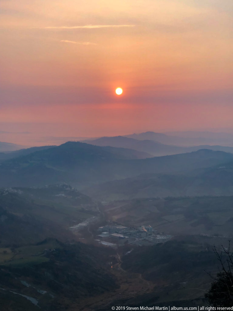 Orange sun setting above layered dark mountain ridges beneath a pink and violet sky.