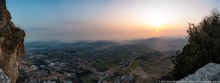 Low sun over hazy valleys, hills, roads, and settlements viewed from a rocky high point on Monte Titano.