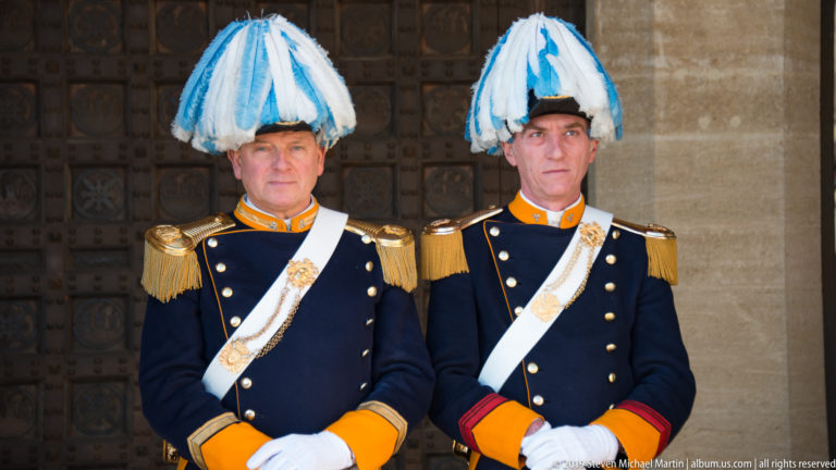 Two San Marino ceremonial guards pose frontally in dark uniforms with gold epaulettes, white belts, gloves, and blue-white plumed hats.