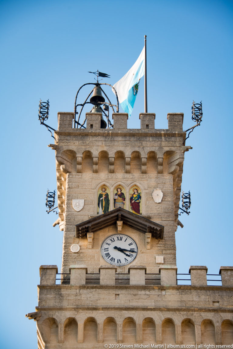 Upper Palazzo Pubblico clock tower with battlements, San Marino flag, three colored panels, clock face, and rooftop metalwork.
