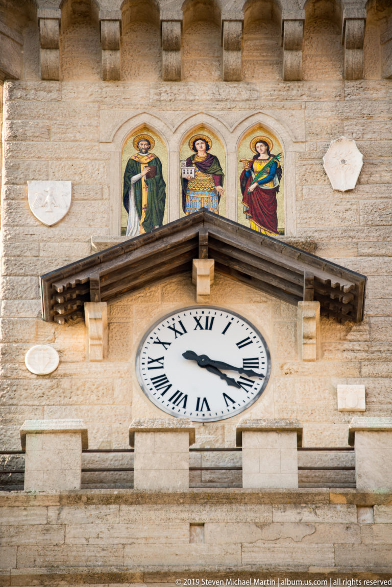Roman-numeral clock beneath three painted arched panels and a small timber canopy on the Palazzo Pubblico tower.