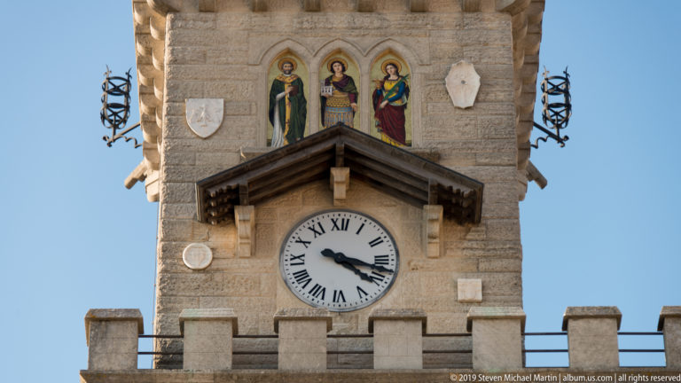 Vertical view of Palazzo Pubblico tower showing battlements, three painted figures, wooden clock canopy, Roman-numeral clock, and side metalwork.