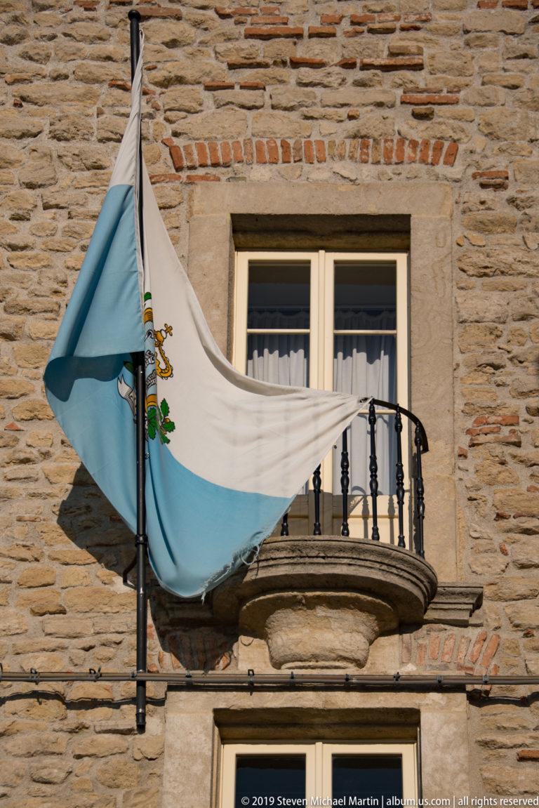 Blue-and-white San Marino flag draped over a small wrought-iron balcony on a sunlit stone building.