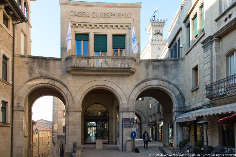 Three stone arches beneath a Cassa di Risparmio façade, with pedestrians and a crenellated tower visible farther along the street.