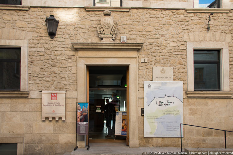 Stone entrance to Museo di Stato with carved crest, museum signs, exhibition posters, and visitors entering through the open doorway.
