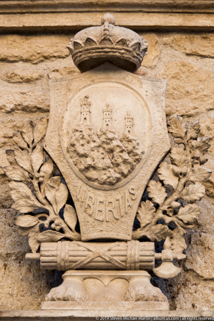 Carved stone emblem with three towers, crown, leafy branches, and the word LIBERTAS on a weathered wall.
