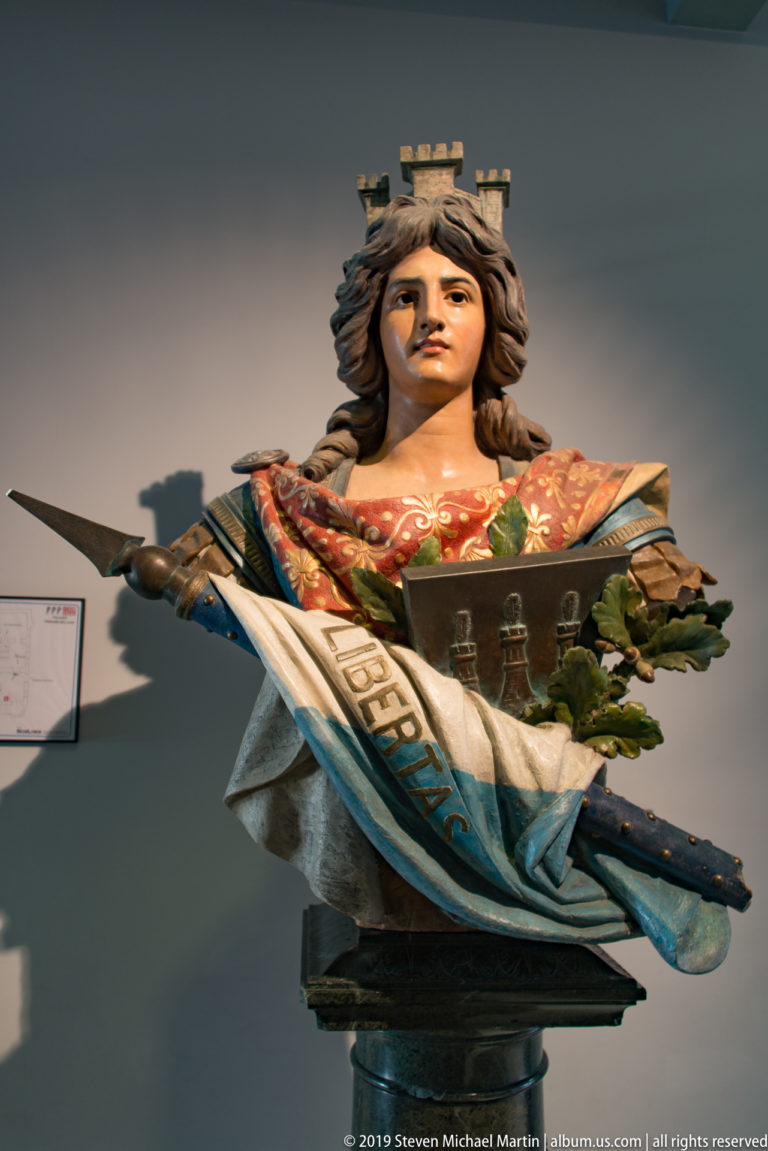 Polychrome crowned female bust wearing a sash marked LIBERTAS, with symbolic objects and foliage, displayed against a museum wall.