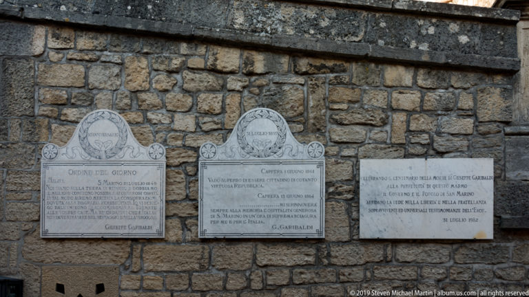 Three inscribed marble commemorative plaques mounted side by side on a rough stone wall, including references to Garibaldi.