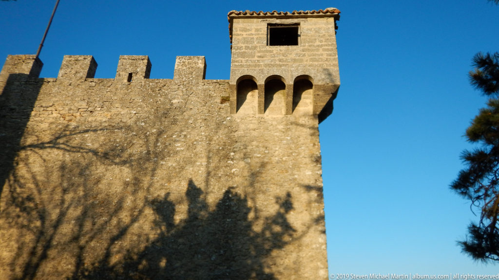 Square stone fortification tower with corbelled openings above a crenellated wall, crossed by dark tree shadows.