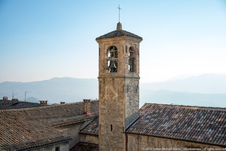 Slender stone bell tower with visible bells, tiled pyramidal roof, and cross rising above terracotta rooftops and distant mountains.