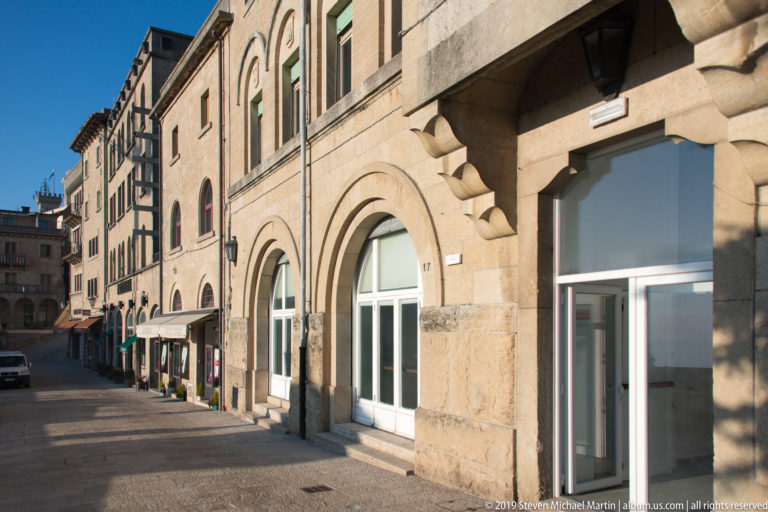 Quiet San Marino street lined with pale stone buildings, round-arched storefronts, shuttered windows, and strong sunlight.