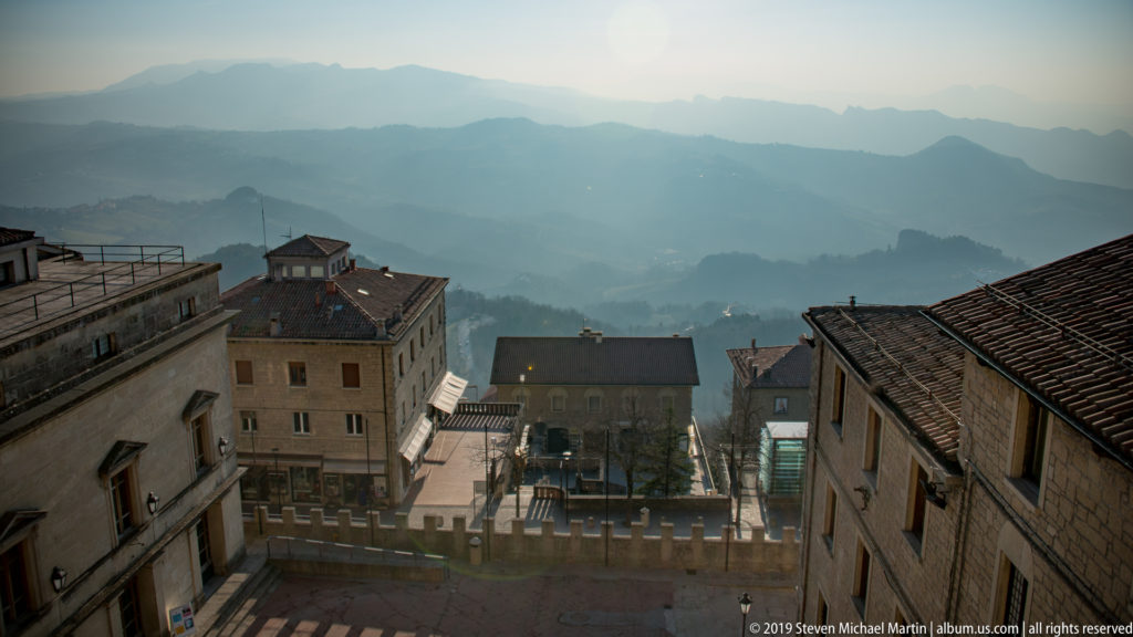 Elevated view over San Marino rooftops and a crenellated parapet toward multiple hazy mountain ridges in the distance.
