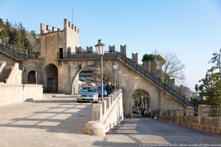 Stone arch, square tower, and crenellated stairway beside a paved route with pedestrians and a vehicle.