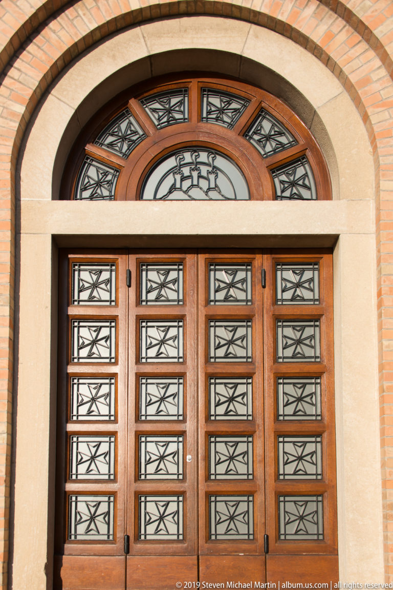 Arched doorway with wooden double doors, geometric metal grilles, semicircular fanlight, and alternating brick-and-stone surround.