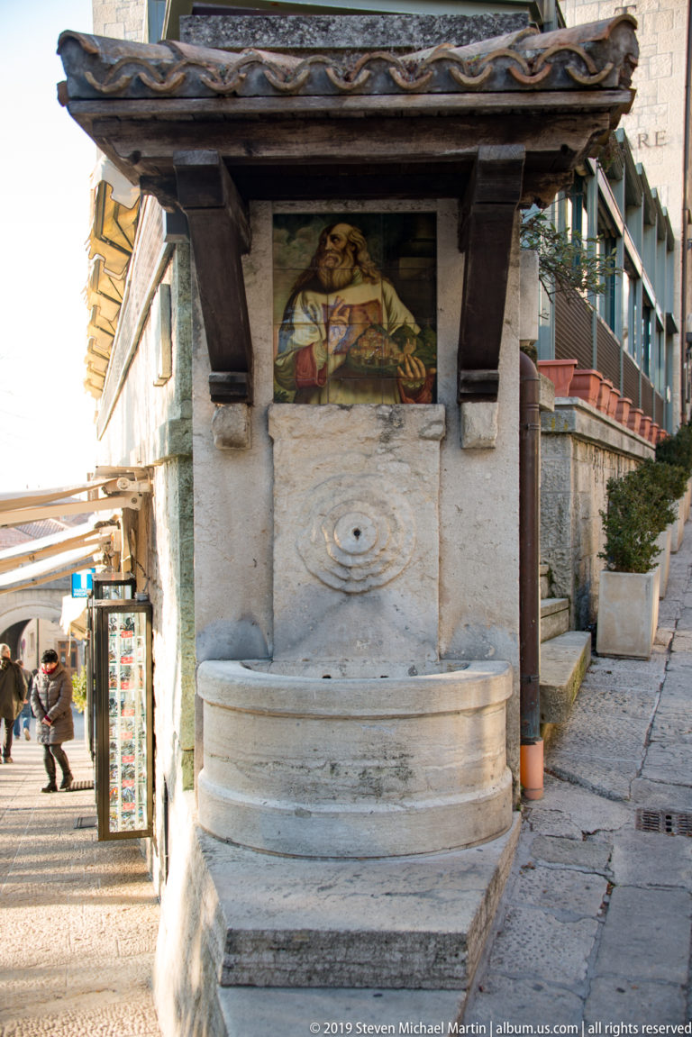 Stone wall fountain with round basin, carved rosette, religious mosaic, and small tiled canopy on a narrow street.