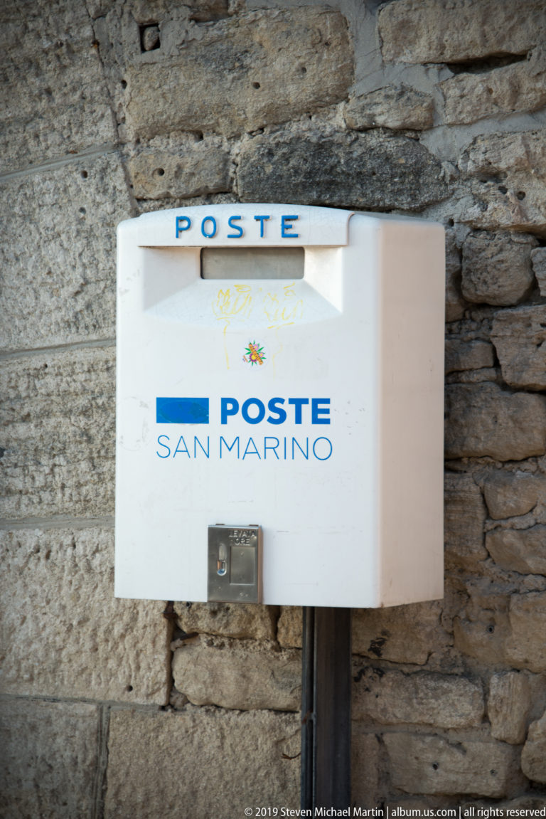 White Poste San Marino mailbox with blue lettering mounted on a rough gray stone wall.
