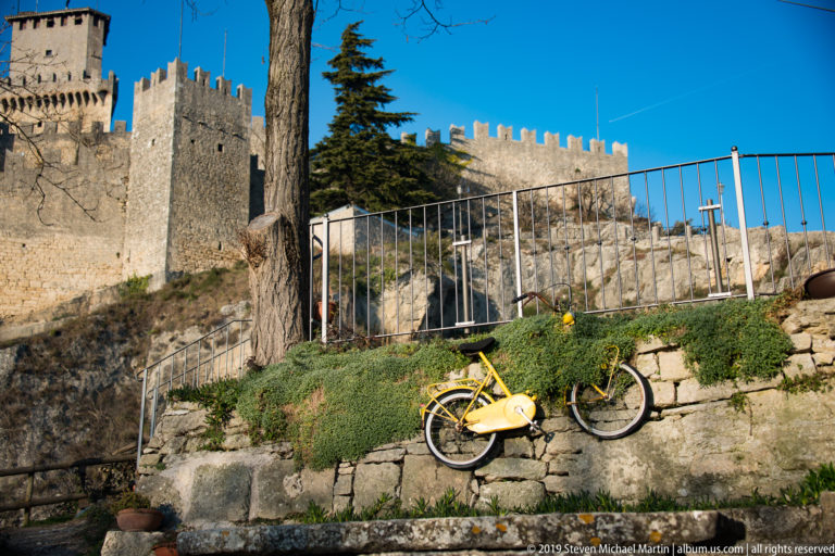 Yellow bicycle leaning below a stone terrace and railing, with crenellated fortification walls rising behind.