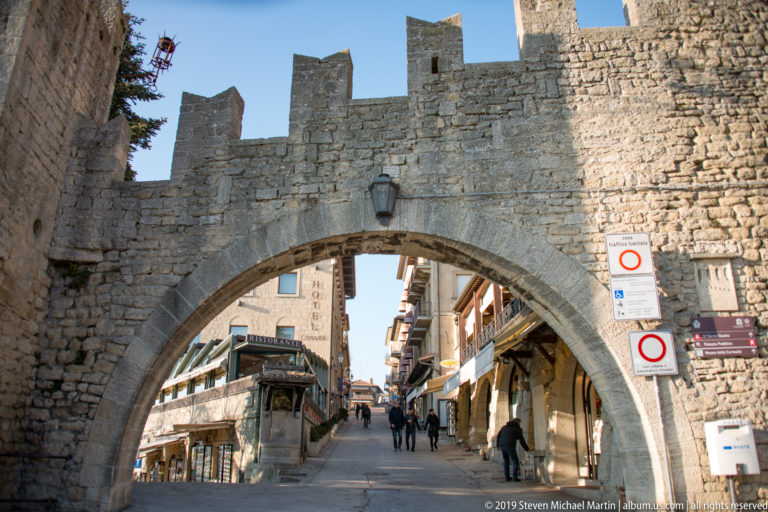Crenellated stone gate with a large round arch framing a narrow uphill street, shops, signs, and pedestrians.