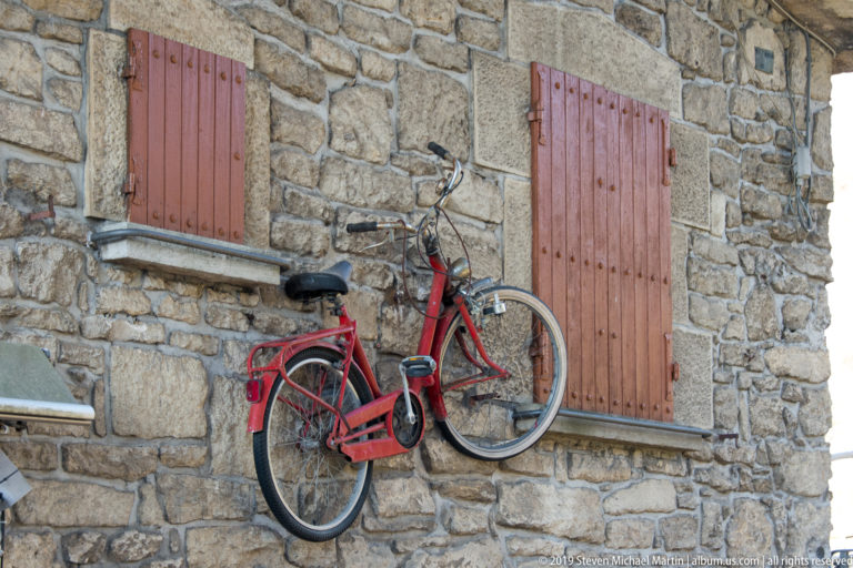 Red bicycle mounted on a rough stone wall between two closed reddish wooden shutters.