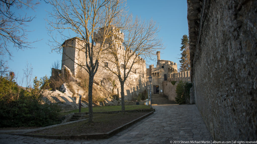 Stone path beside a high wall leading uphill toward crenellated fortress towers among bare trees and rock.