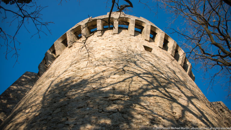 Low-angle view up a cylindrical stone defensive tower with projecting crenellations and tree shadows.